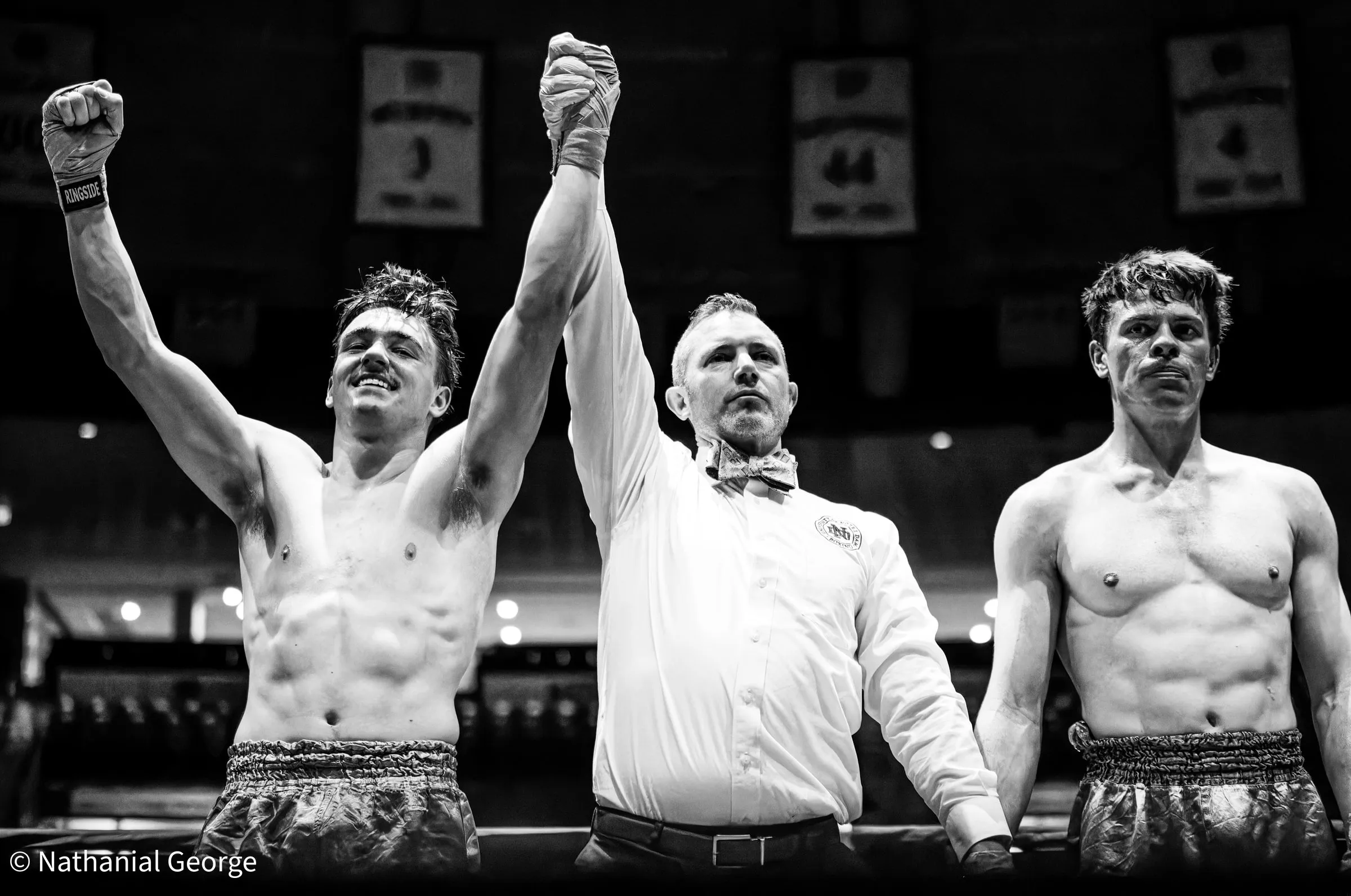 In black and white, a referee in a bow tie lifts the arm of a boxer who grins with his other fist in the air, while his opponent stands to the right under banners hanging from the rafters.