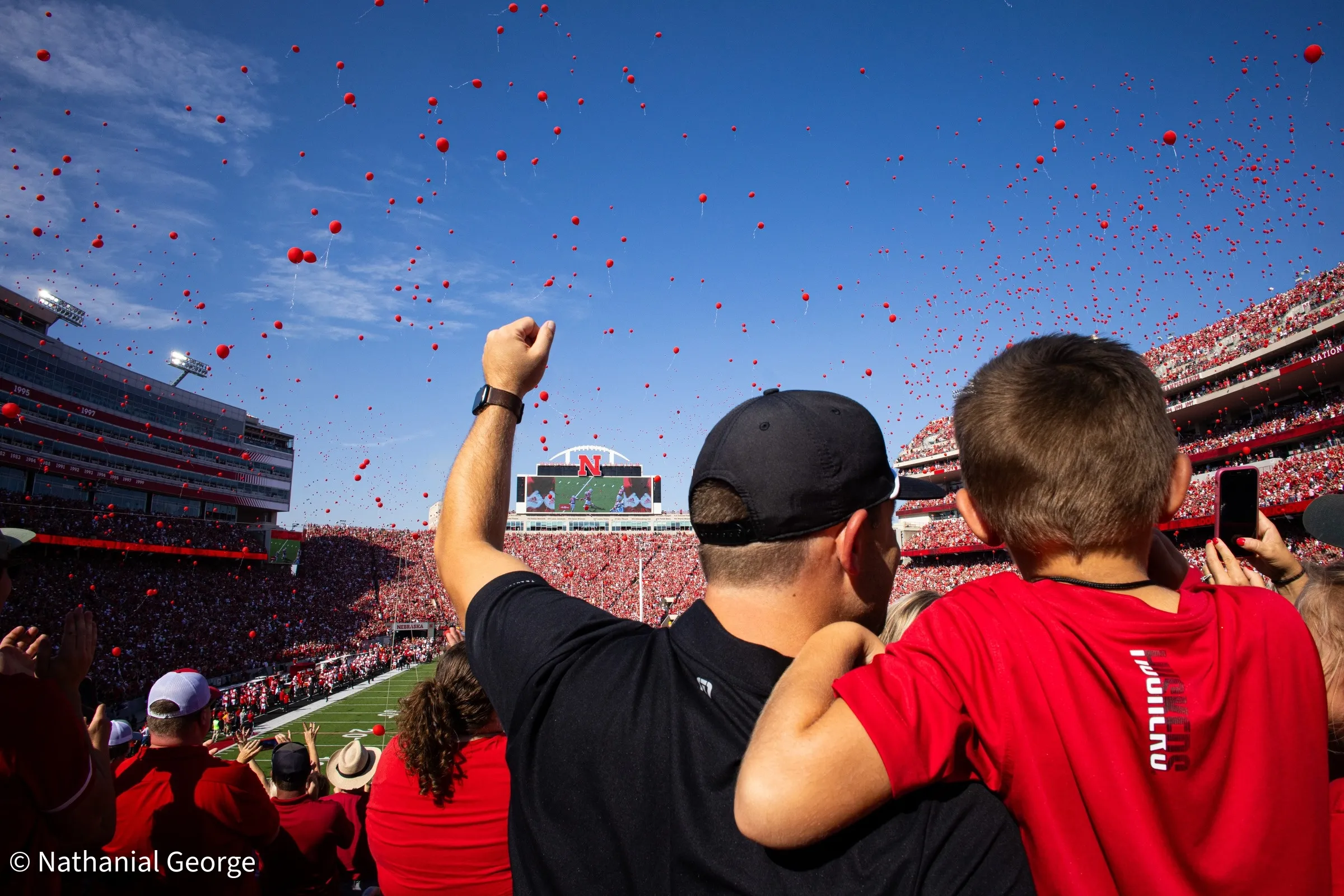 A fan in a black cap raises a fist beside a boy in a red shirt as a sky full of red balloons rises over the packed stadium.